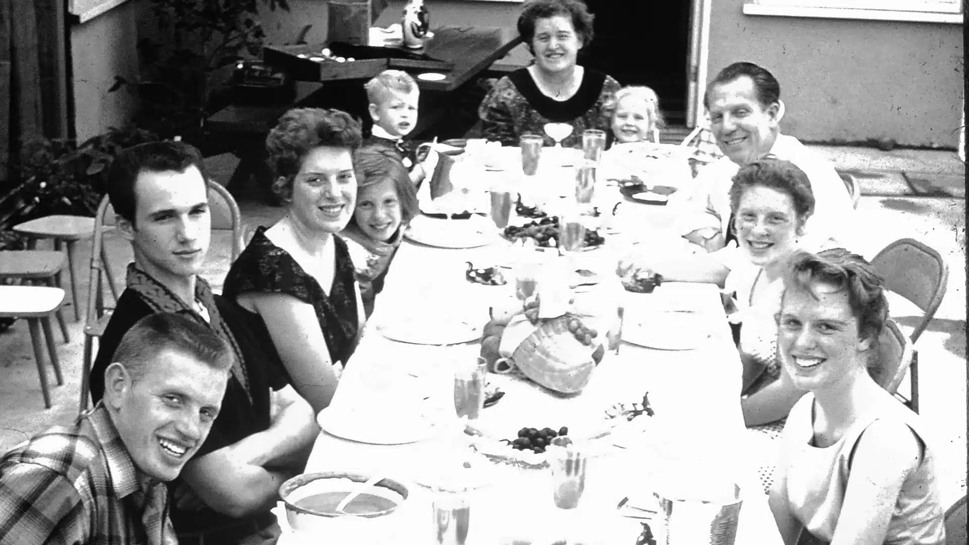 A group of smiling people gathered around a long table set for a meal outdoors.