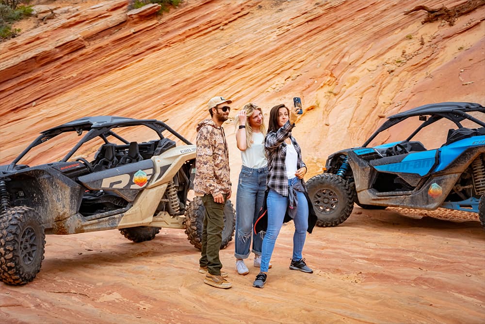 Three people take a selfie in front of off-road vehicles on a sandy landscape.
