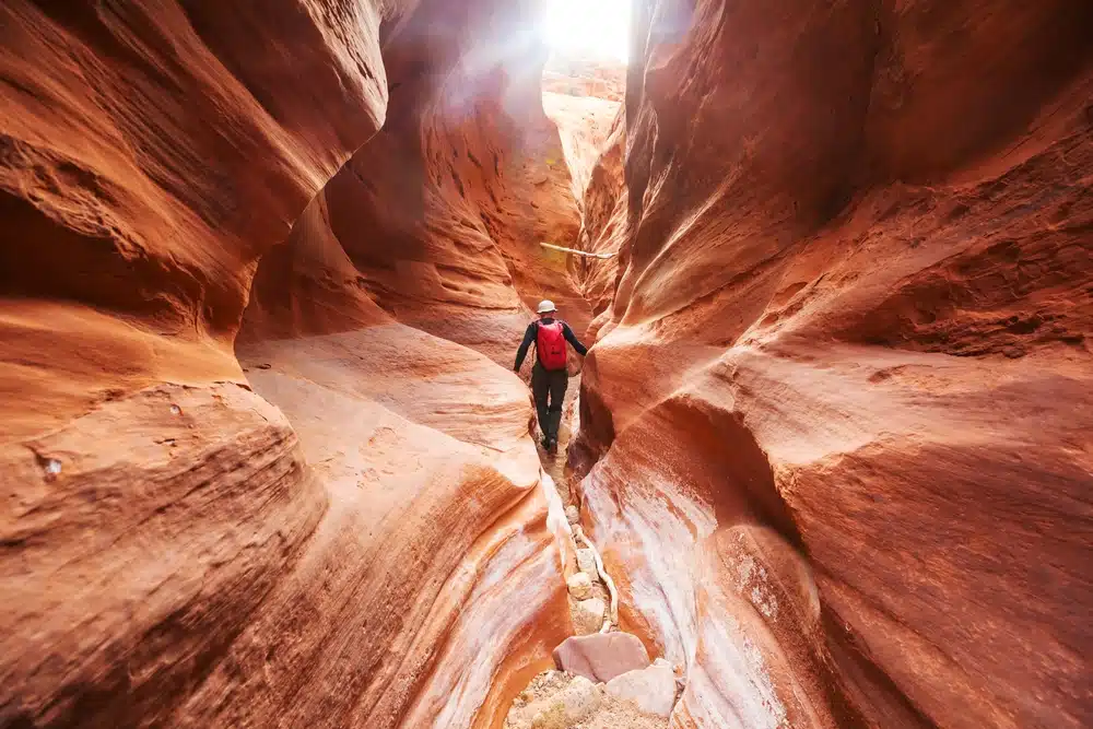 A hiker navigating a narrow, sunlit canyon with red rock walls.