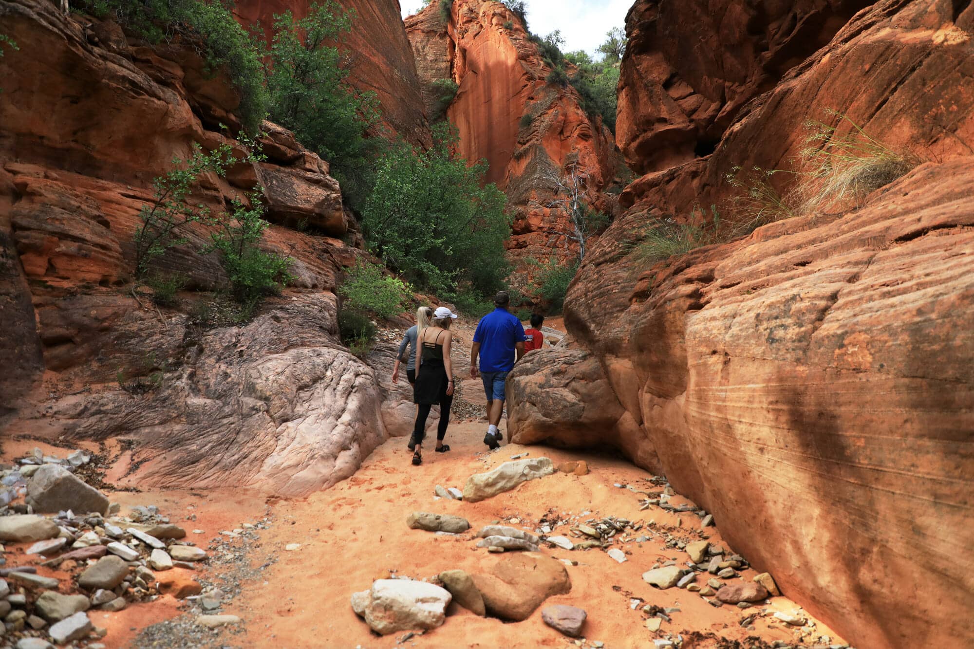 Two hikers walking along a sandy trail in a narrow, red rock canyon.