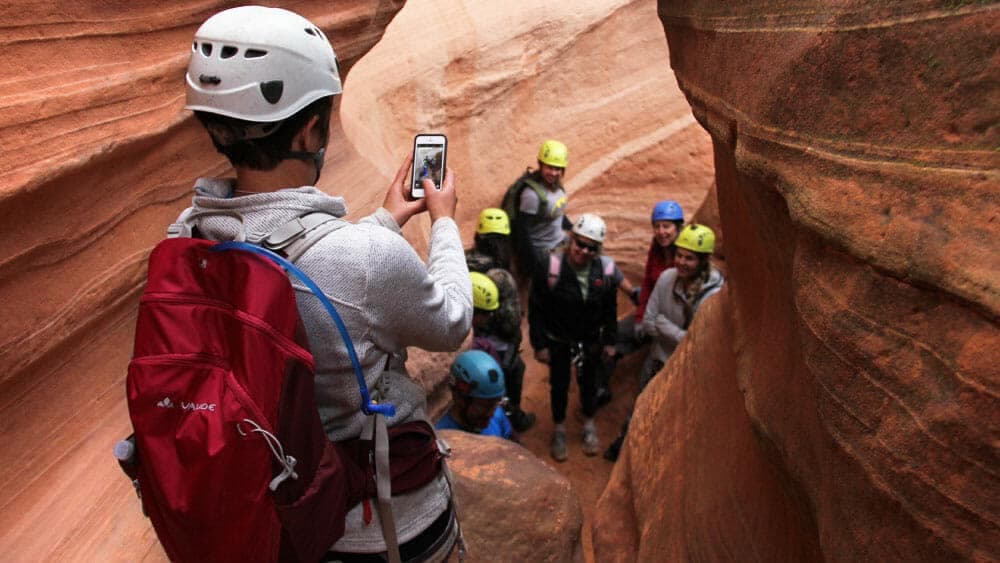 A person in a helmet takes a photo of a group of individuals in helmets standing in a narrow canyon.
