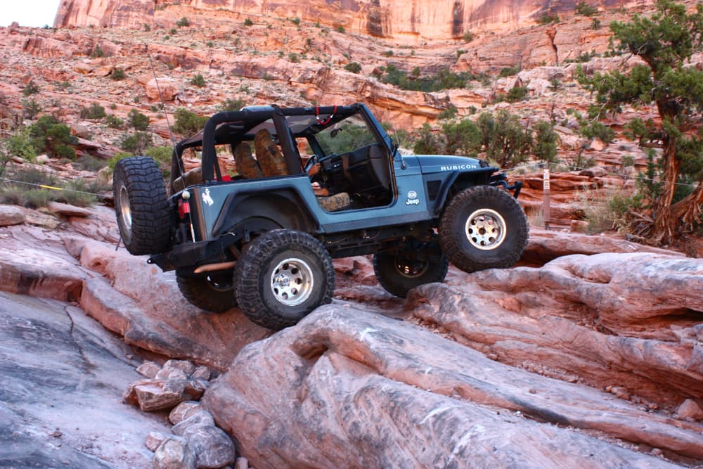 A blue off-road vehicle climbs over rocky terrain in a desert landscape.