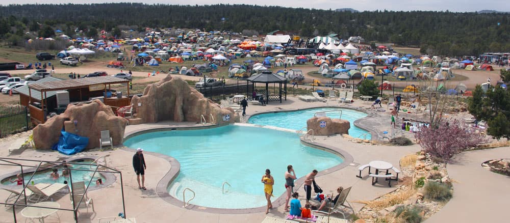 A view of a swimming pool area with people relaxing, surrounded by a large camping site filled with colorful tents in the background.