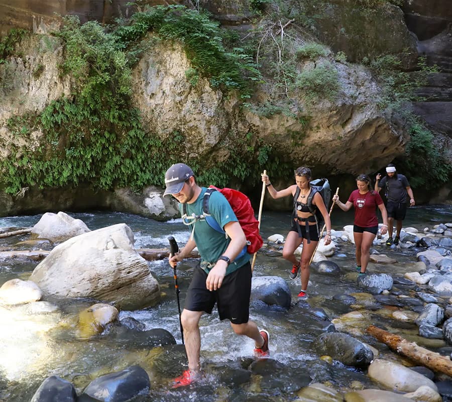 A group of four hikers wades through a rocky river surrounded by lush greenery.