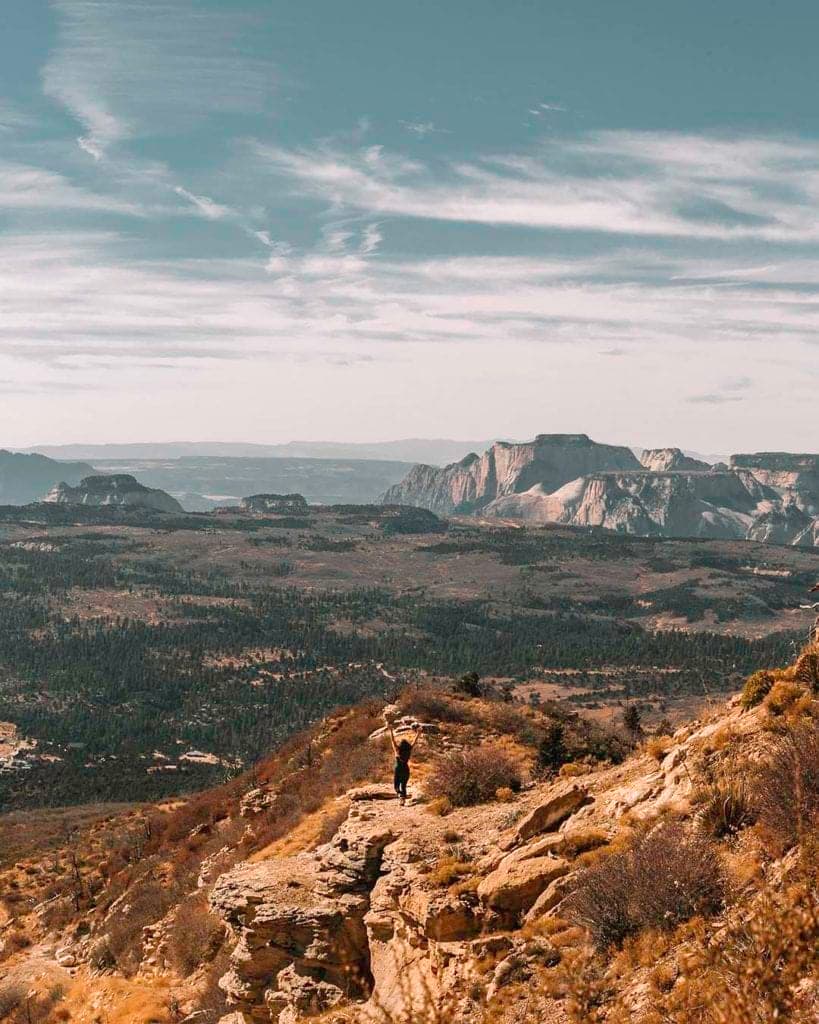 A lone hiker stands on a rocky path overlooking a vast mountainous landscape under a blue sky.