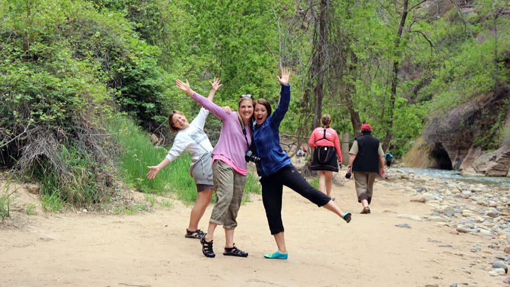 Three women playfully pose by a creek while others walk in the background, surrounded by lush greenery.