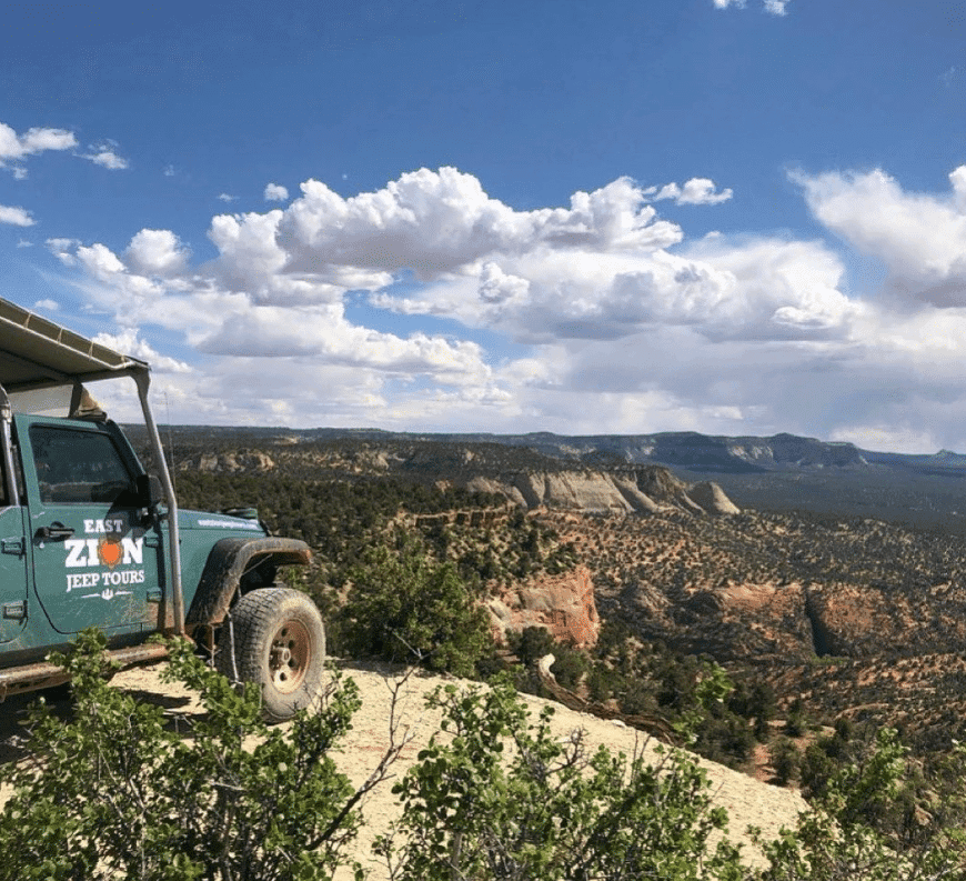 A green jeep rests on a cliff overlooking a vast desert landscape under a cloudy sky.