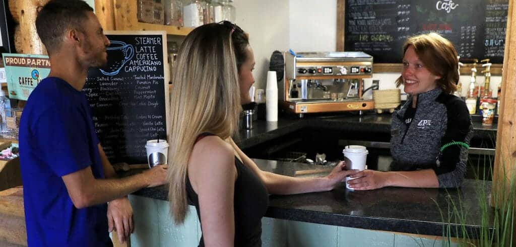 A barista serves coffee to a customer while another customer engages in conversation at a café.