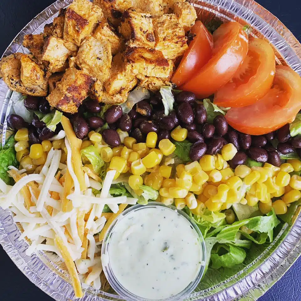 A colorful salad with grilled chicken, black beans, corn, lettuce, tomatoes, shredded cheese, and a side of dressing.