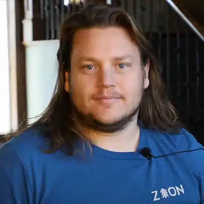 A man with long hair wearing a blue shirt stands in a well-lit setting.