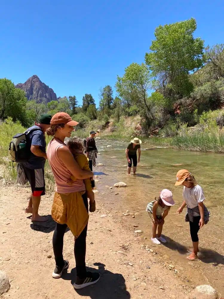 A group of children and adults are playing by a shallow river in a scenic outdoor setting.