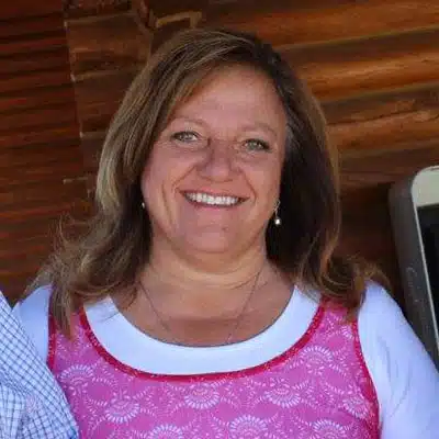 A smiling woman with long brown hair wearing a pink blouse stands against a wooden background.
