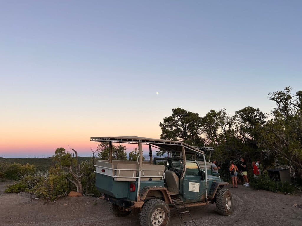 A jeep parked on a dirt trail with people gathered nearby under a colorful sunset sky.