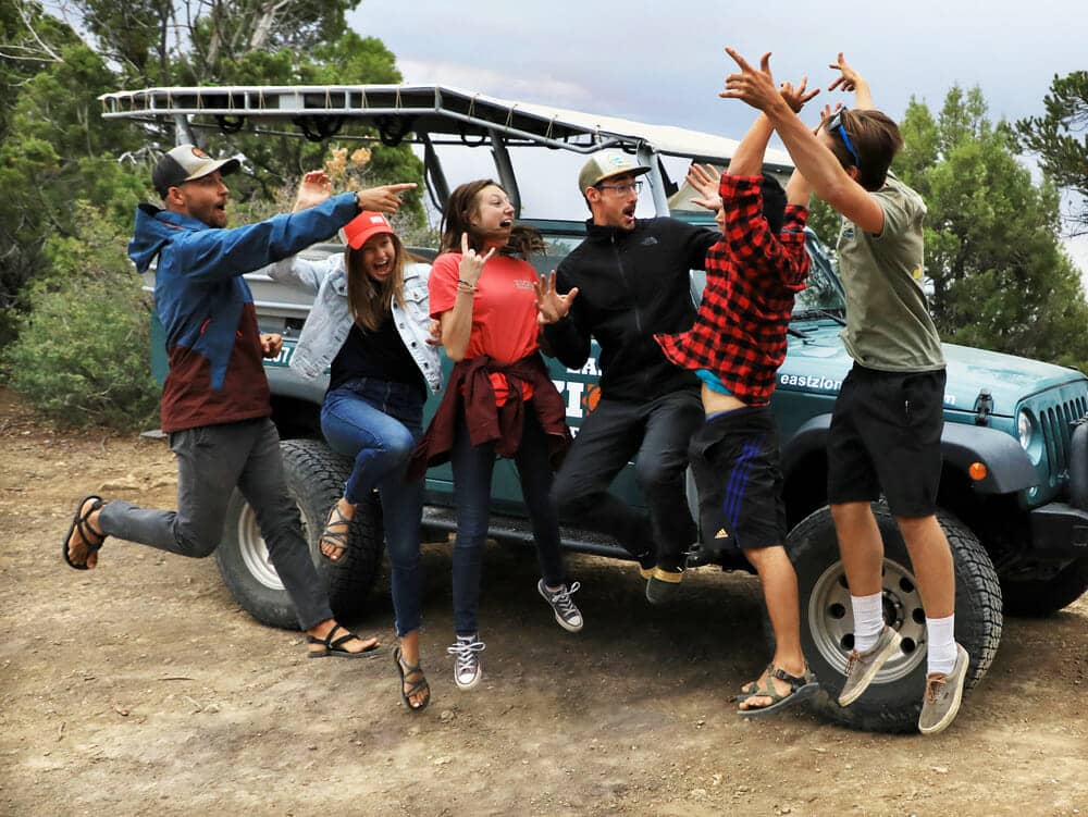 A group of six friends joyfully jumping and posing near a parked Jeep in a forested area.