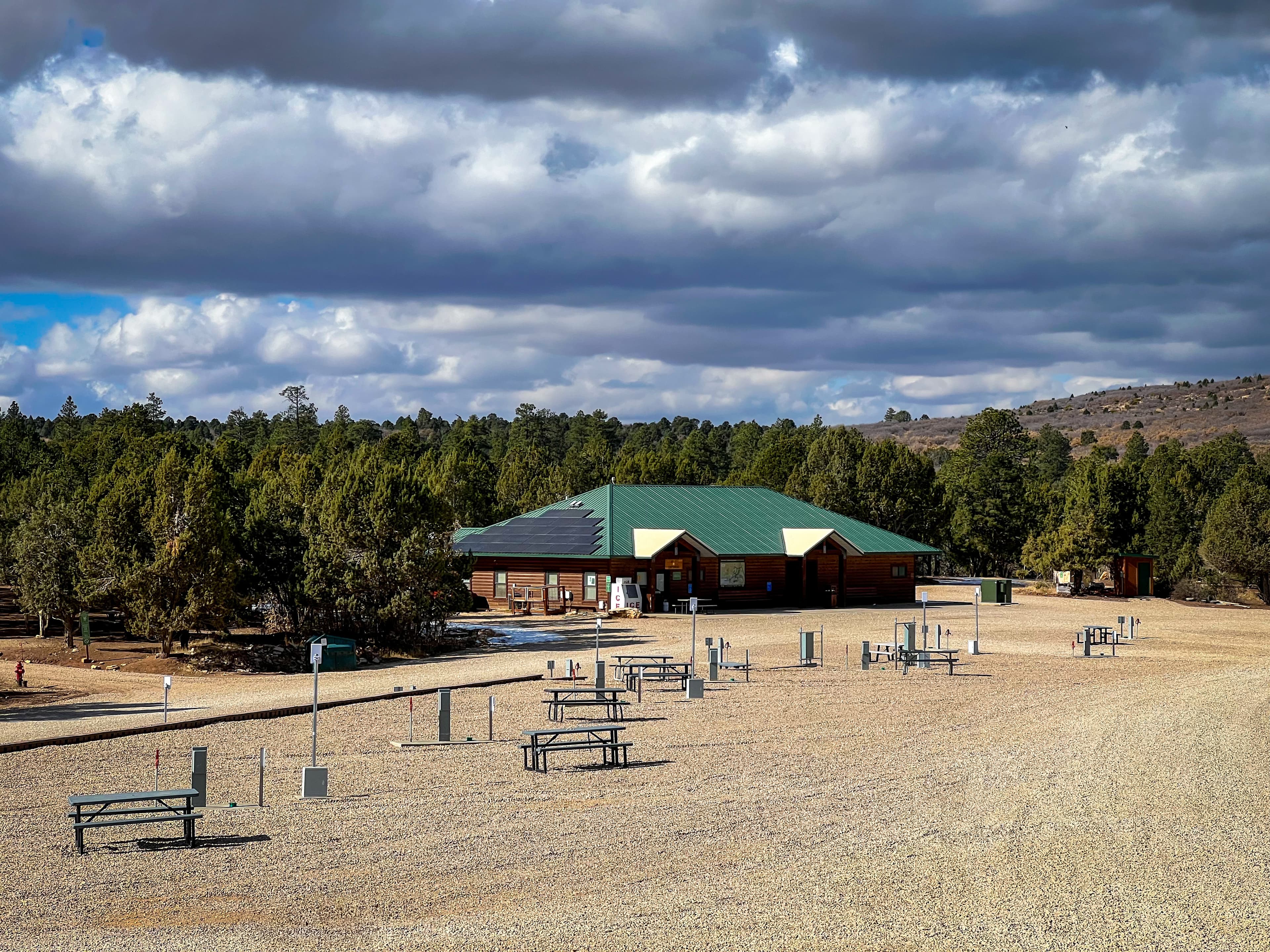 A rustic building with a green roof surrounded by picnic tables and trees under a cloudy sky.