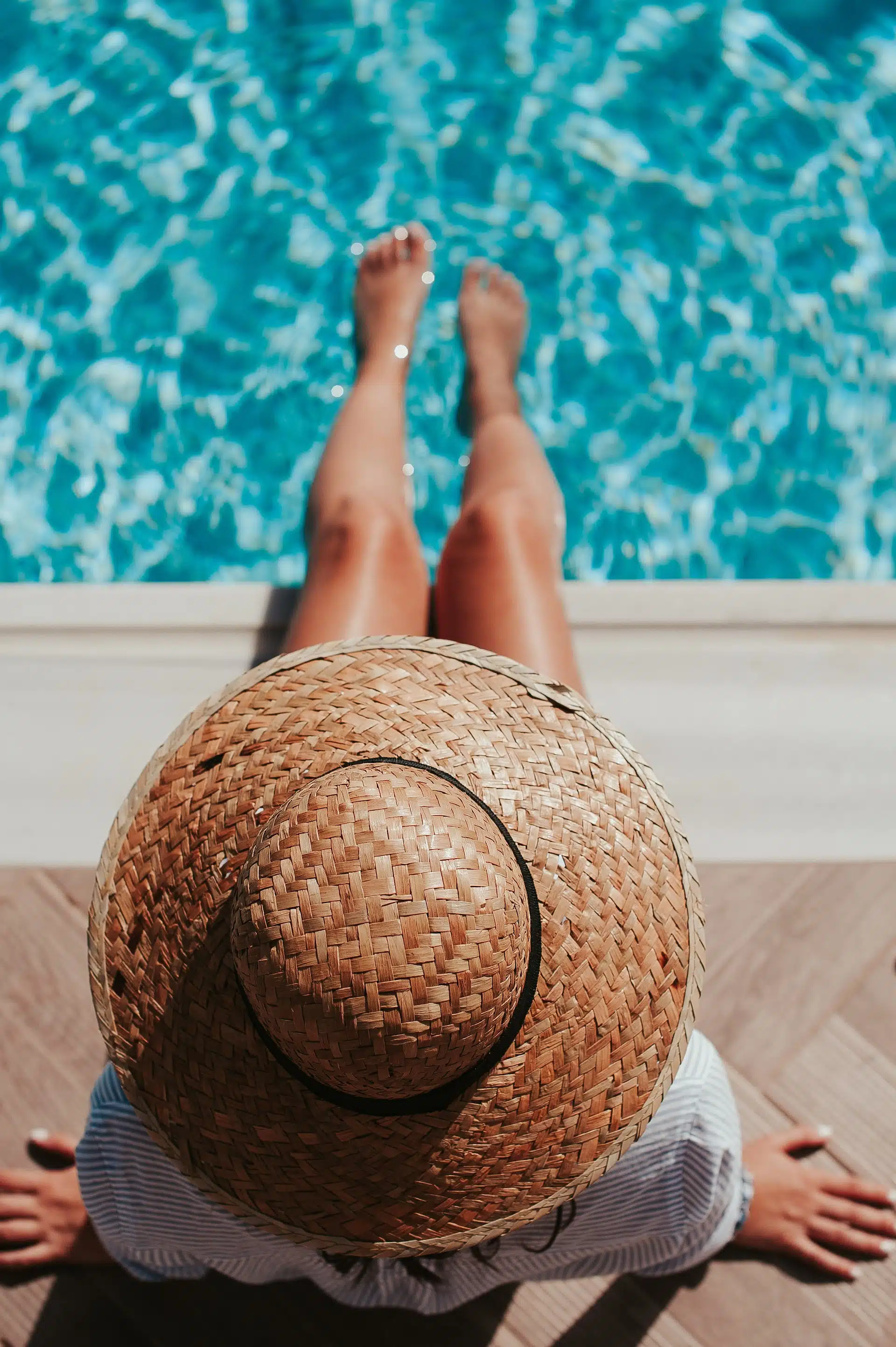 A person wearing a straw hat sits by the edge of a pool, with their bare feet in the water.