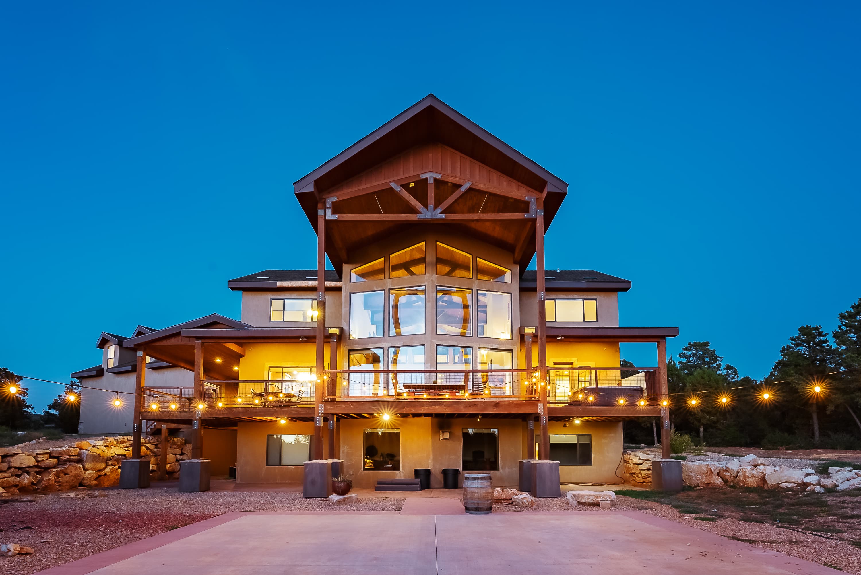 A modern multi-story house with large windows, surrounded by trees, illuminated at dusk.