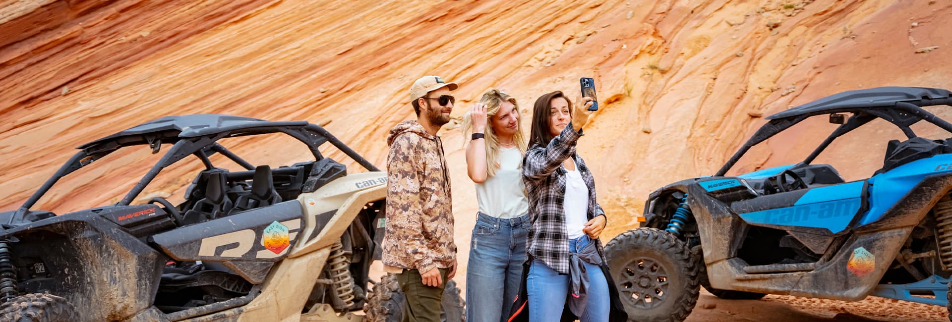 Three friends pose for a selfie next to off-road vehicles in a desert landscape.