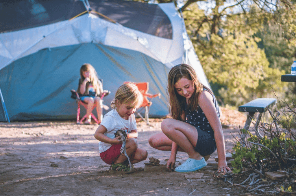 Two children play in the sand near a camping tent while another child sits in a chair in the background.