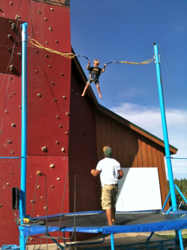 A child swings high above a trampoline while an adult watches attentively in a sunny outdoor setting.