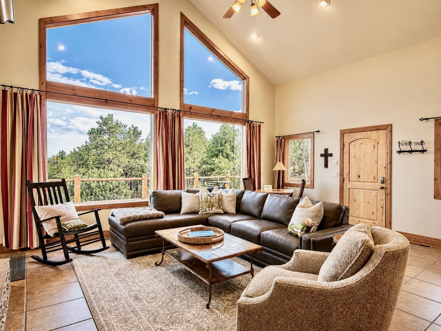 A cozy living room with a large sectional sofa, a coffee table, and tall windows offering views of trees and blue skies.