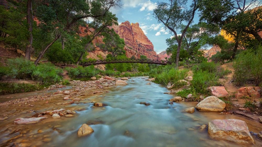 A serene river flows through a lush canyon with rocky cliffs and a wooden bridge in the background.