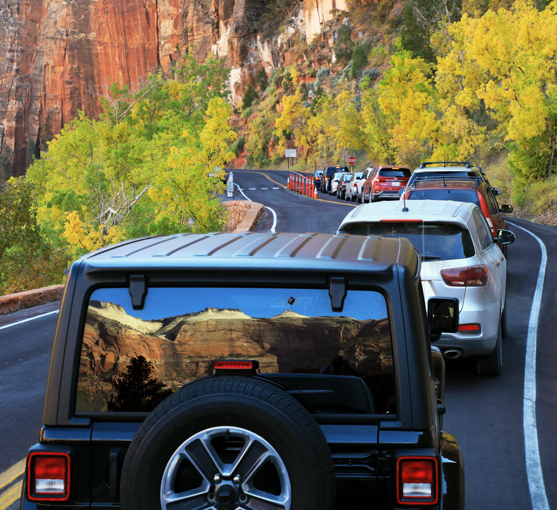 A line of cars drives along a winding road surrounded by autumn-colored trees and cliffs.