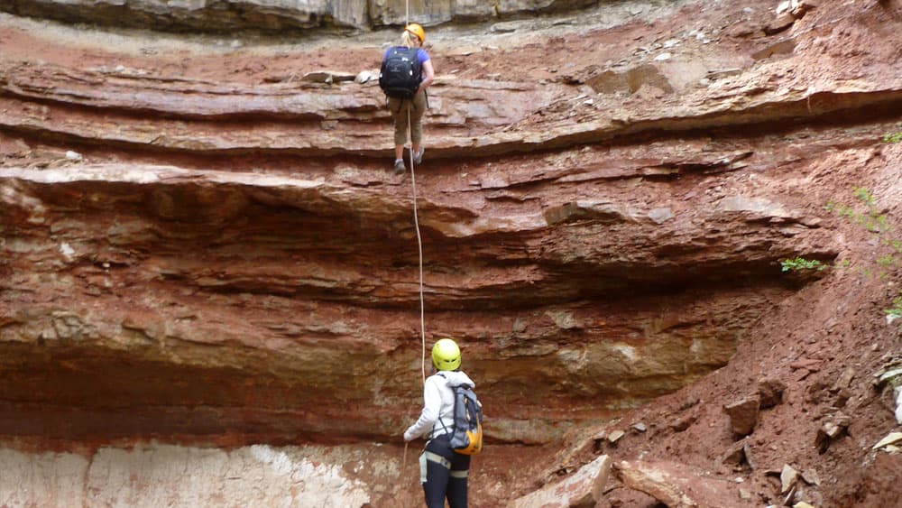 A person climbs a rock face while another person anchors below, both wearing helmets.