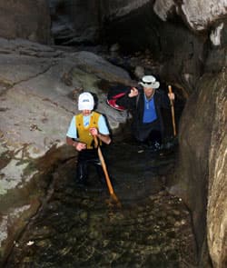Two people are wading through a narrow, rocky creek, using walking sticks for support.