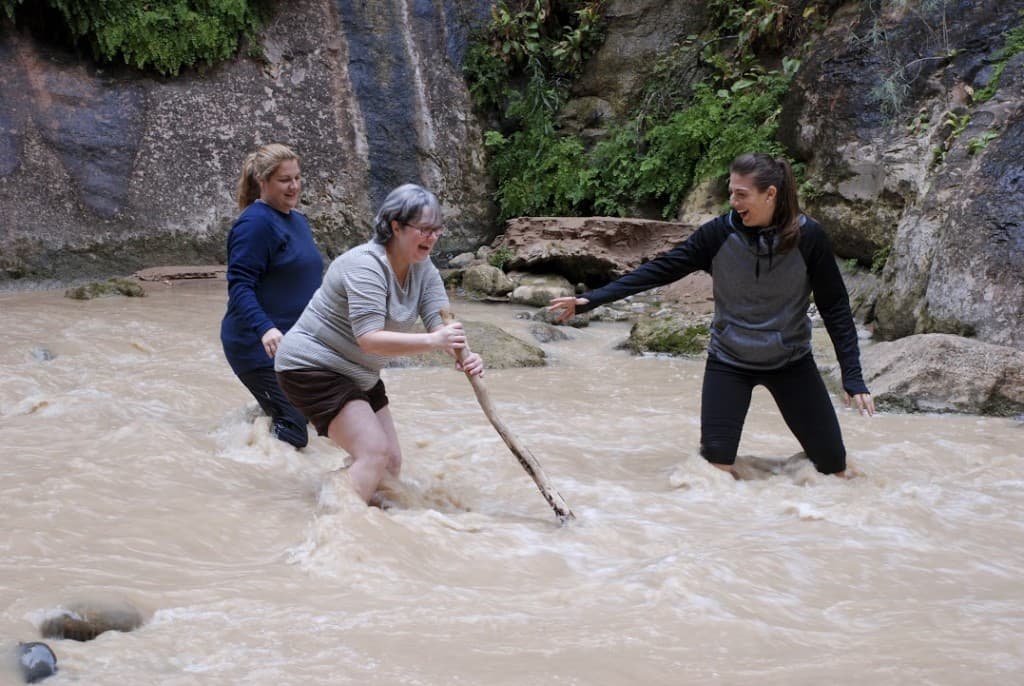 Three women navigate a muddy stream, with one using a stick for support.
