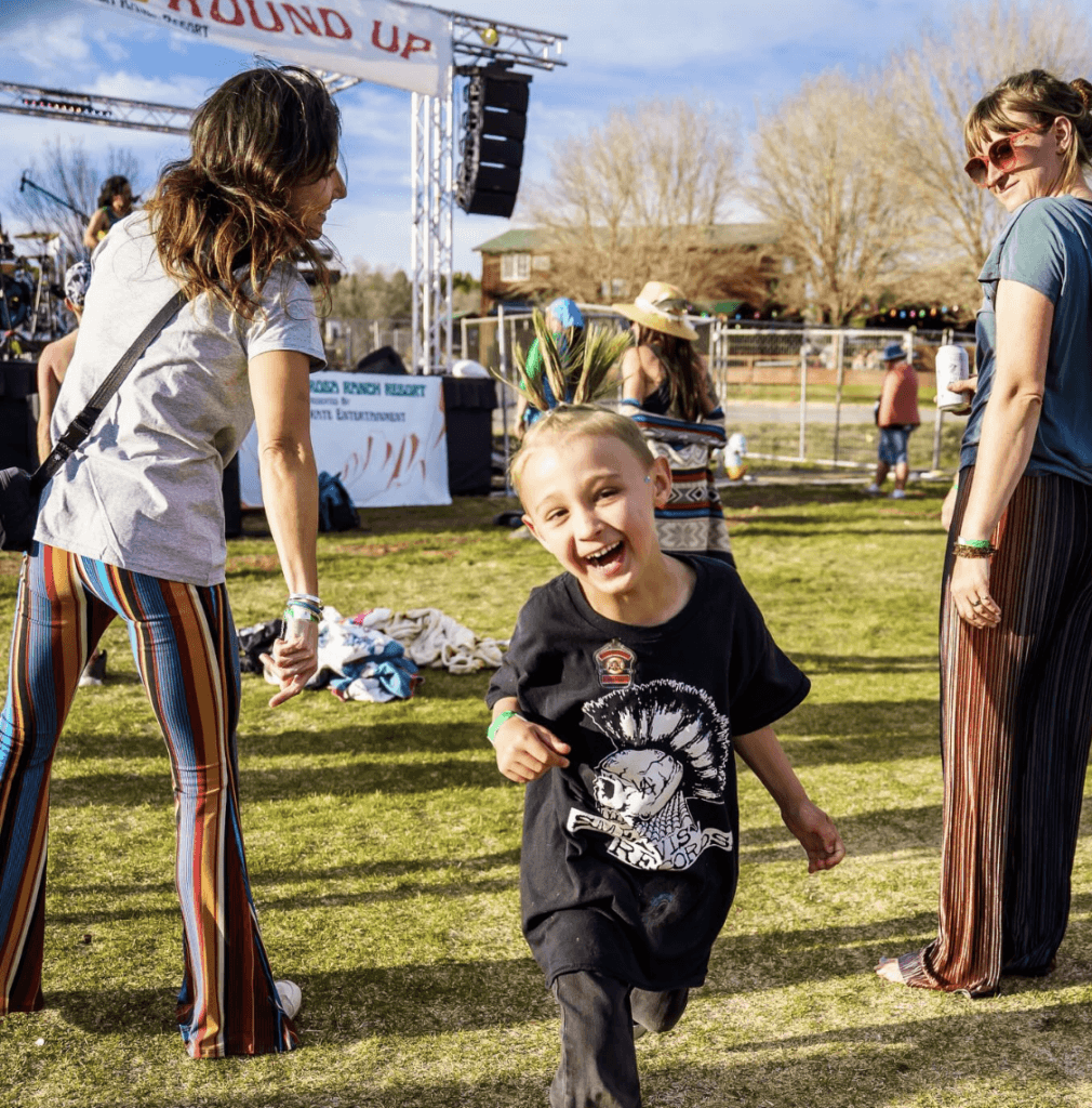A smiling child with a mohawk runs joyfully through a festival, surrounded by adults dancing.