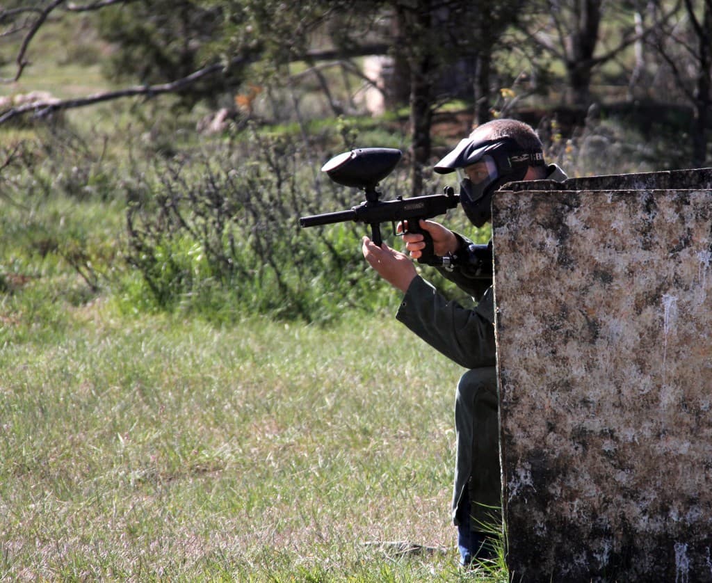 A person in a paintball mask crouches behind a barrier, aiming a paintball gun.