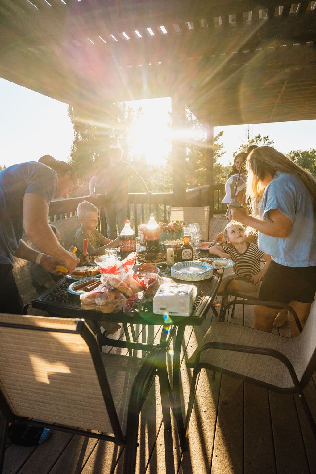 A family gathers around a table decorated with food and drinks during a sunny outdoor barbecue.