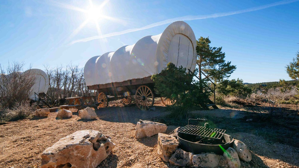 A pair of covered wagons sit under a bright sun in a rugged outdoor setting, near a grill and scattered stones.