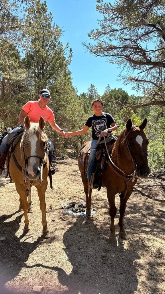 A man and a child on horseback hold hands while riding through a wooded area.