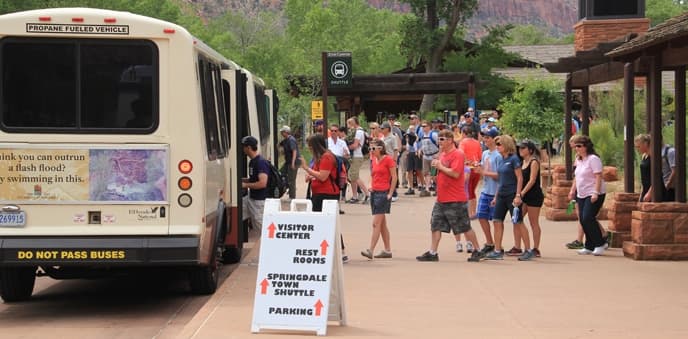 A crowd of people gathers near a shuttle bus and information signs at a visitor center in a scenic park.