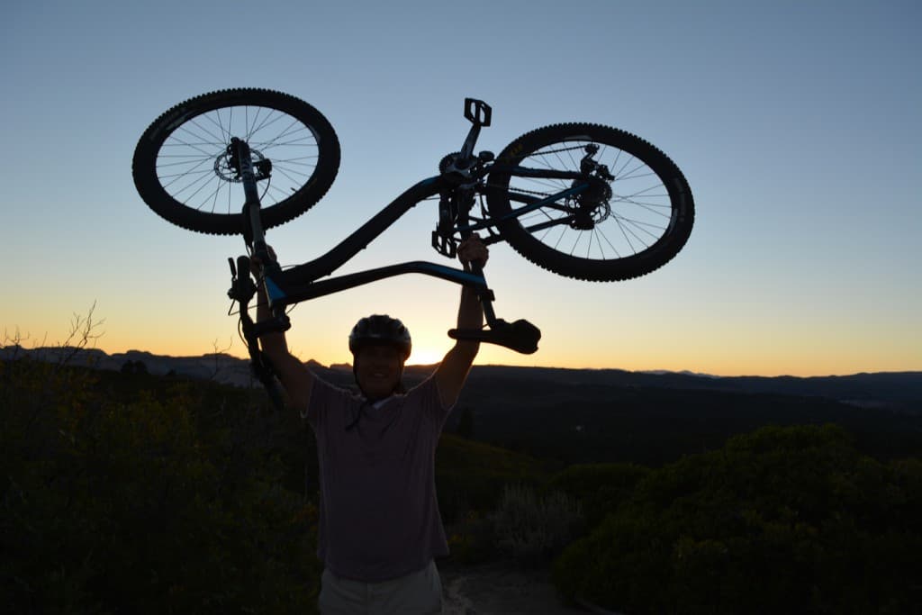 A person wearing a helmet triumphantly raises a mountain bike above their head against a sunset backdrop.