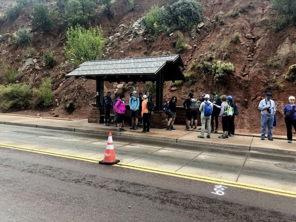 A group of hikers wait at a bus stop, sheltered by a wooden awning, near a red rock hillside.