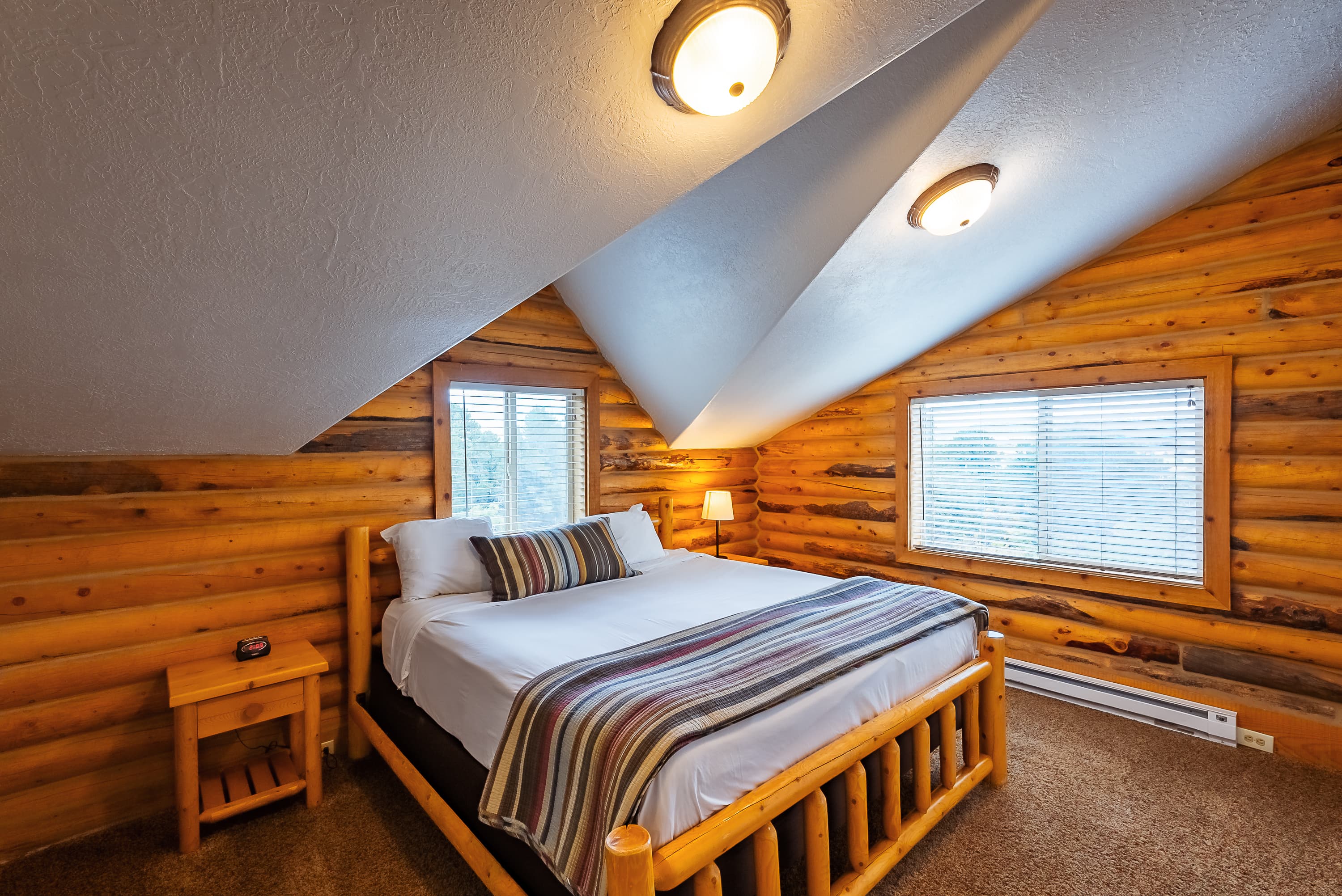 Cozy log cabin bedroom with a wooden bed, striped blanket, and natural light from two windows.