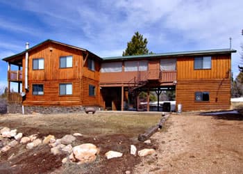 A large wooden cabin with multiple stories and a sloped roof, surrounded by a grassy area and scattered rocks.