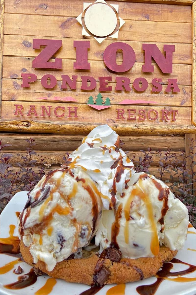 A large cookie topped with scoops of ice cream, whipped cream, and drizzled with chocolate and caramel sauce, in front of a wooden sign for Zion Ponderosa Ranch Resort.