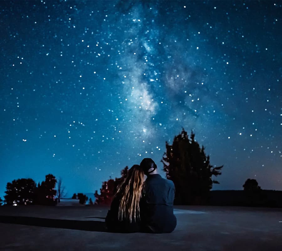 A couple sits together gazing at a starry night sky filled with the Milky Way.