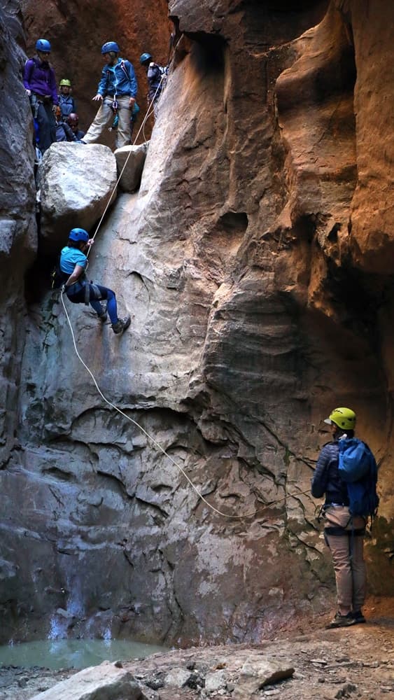 A climber rappels down a rocky canyon while others observe from above.