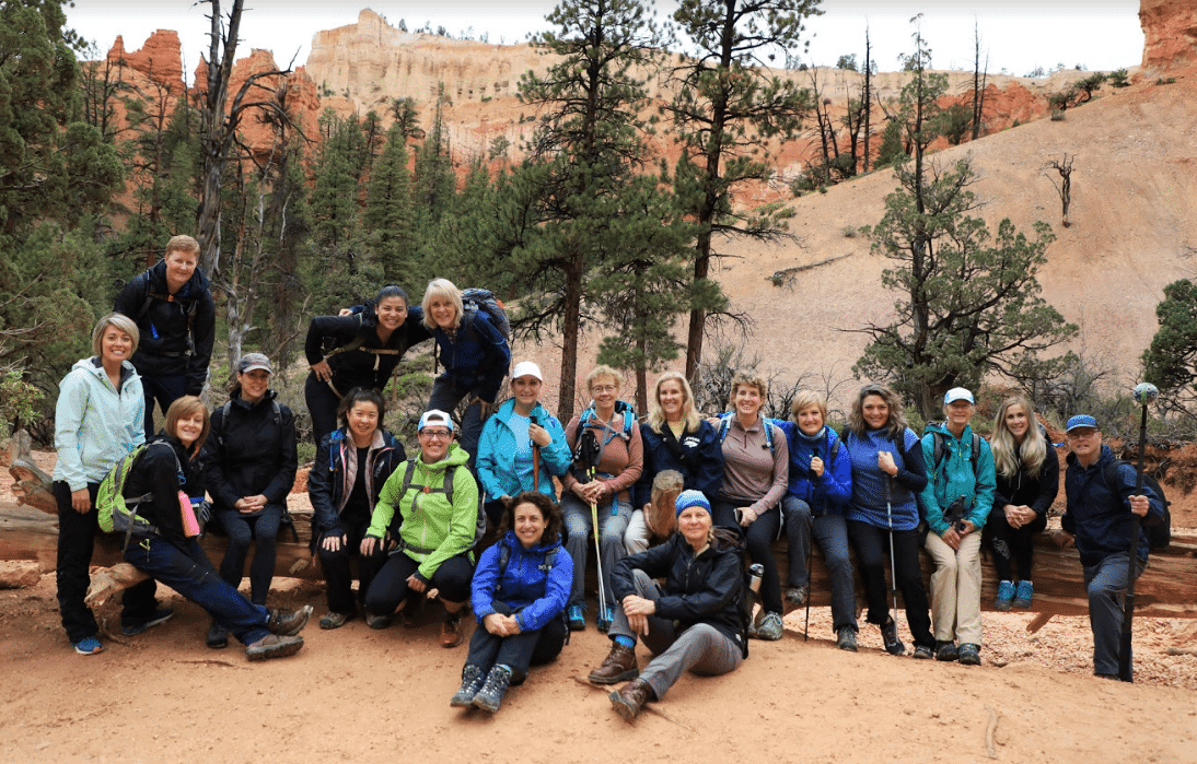 A group of twenty hikers poses together in a scenic outdoor setting surrounded by trees and rock formations.