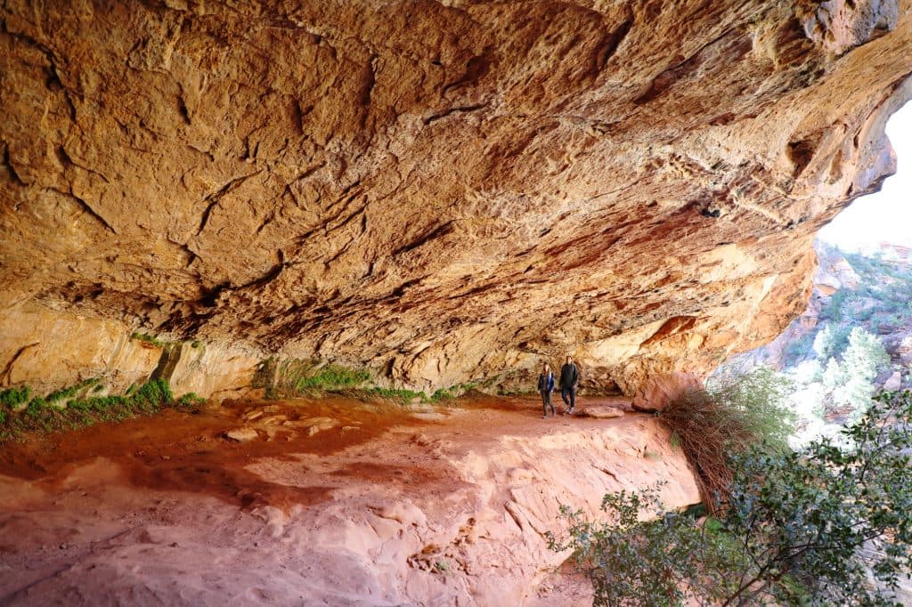 Two hikers explore a sheltered rocky overhang in a desert landscape.