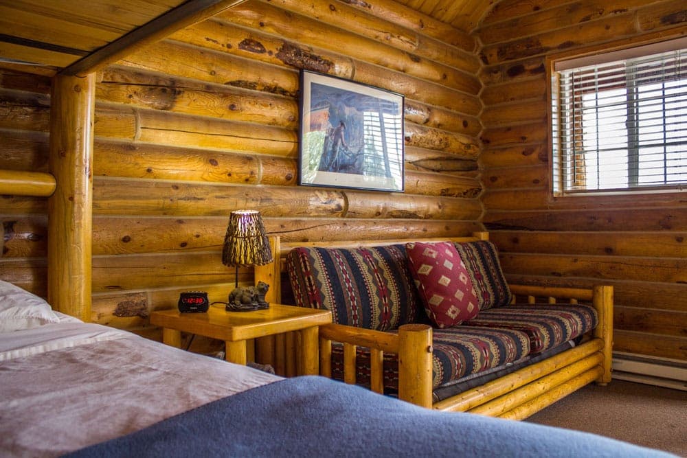 Cozy interior of a log cabin bedroom featuring a bed, wooden sofa, and a small lamp.