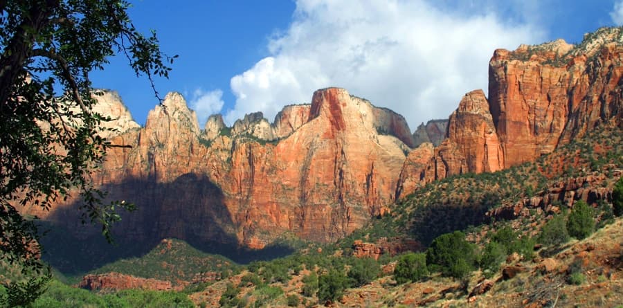 A panoramic view of towering red rock formations against a blue sky with scattered clouds.