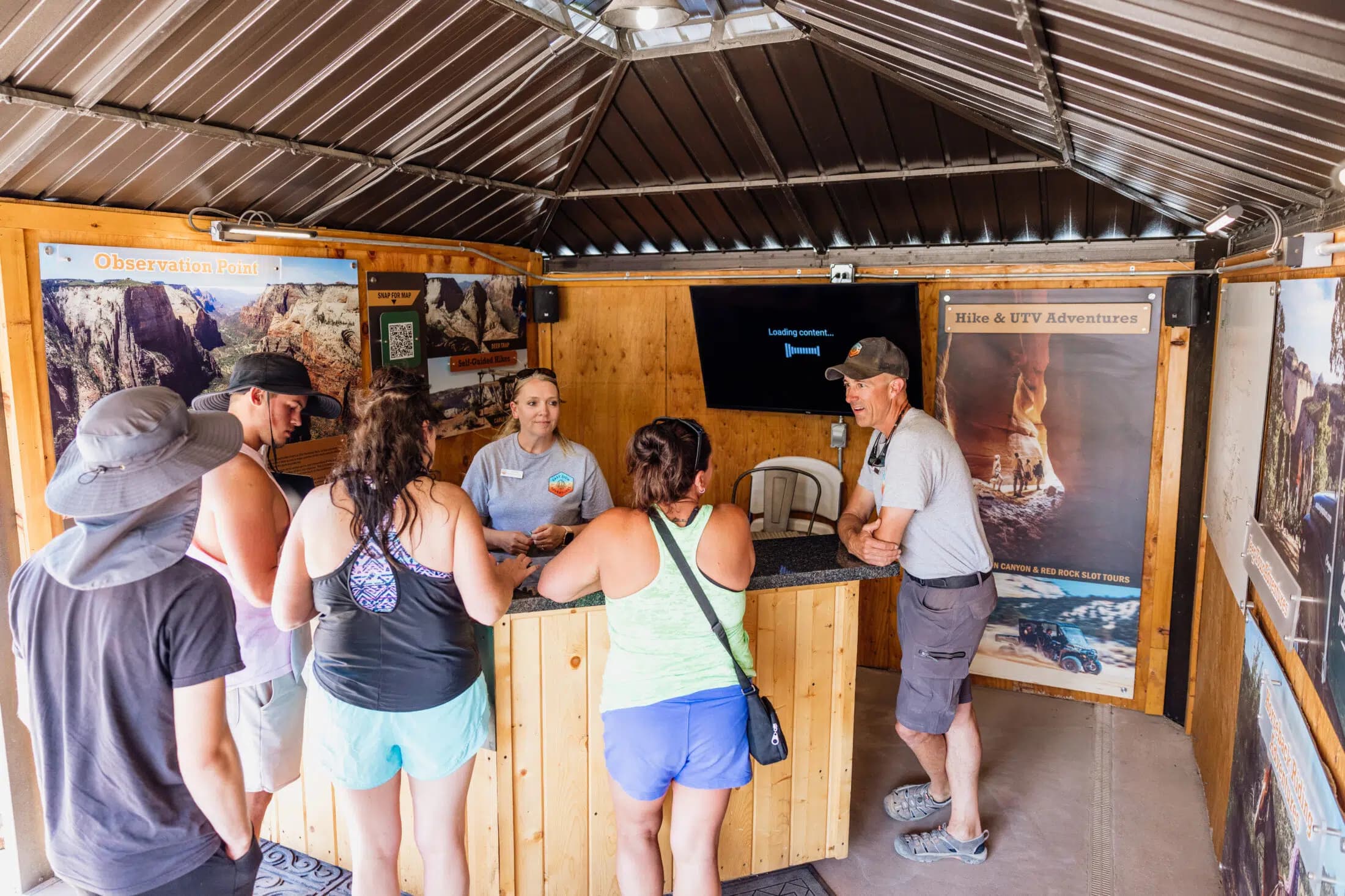 A group of people gathers at a wooden reception desk in an outdoor adventure center, speaking with a staff member.