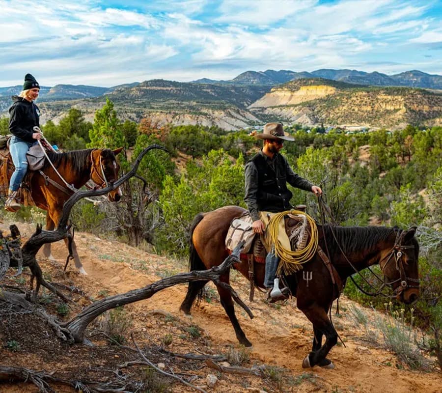 Two riders on horseback navigate a mountainous trail surrounded by greenery.