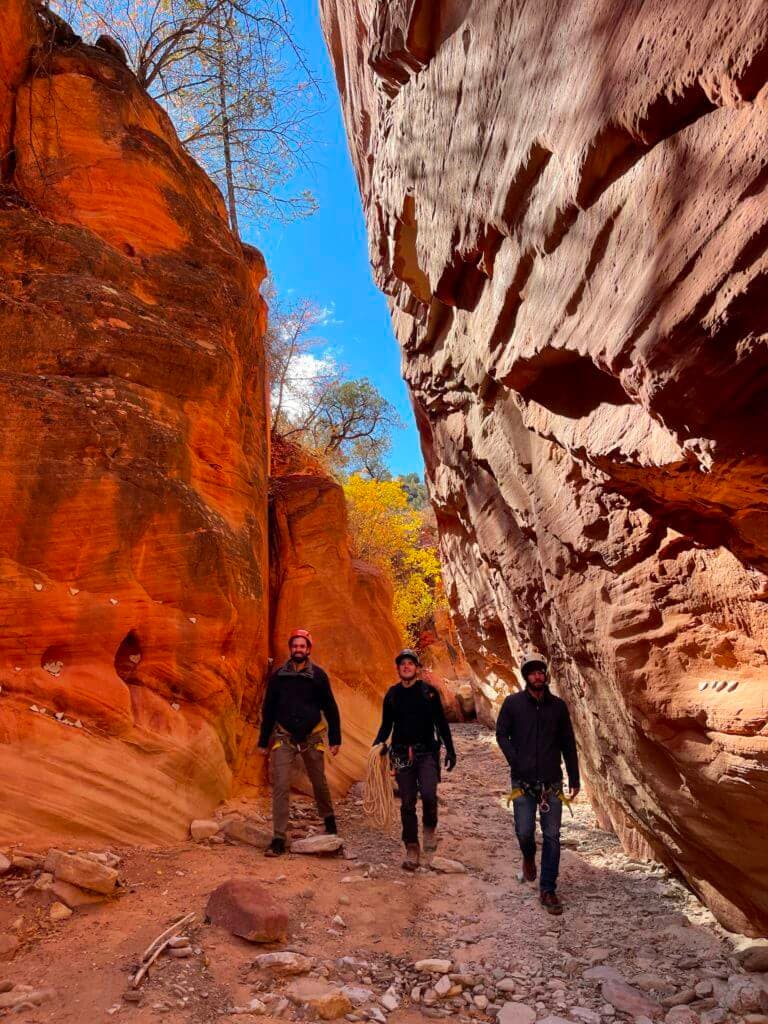Three climbers walk through a narrow, orange canyon surrounded by towering rock walls.